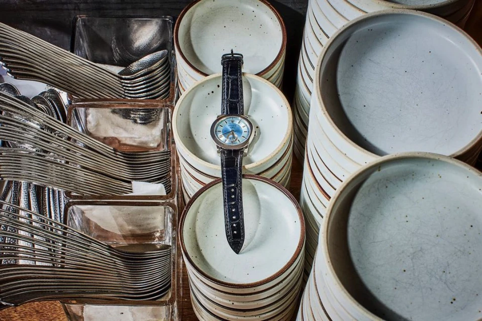 Overhead shot displays a black wristwatch on a stack of white ceramic plates with stacks of plates and silver cutlery surrounding it.