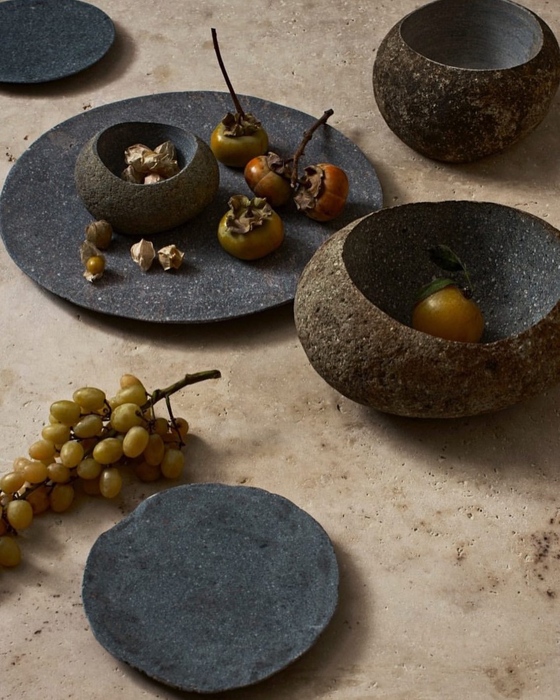 Still life shot of persimmons and grapes on a stone surface, featuring stone bowls and dishes, creating a natural, rustic presentation.