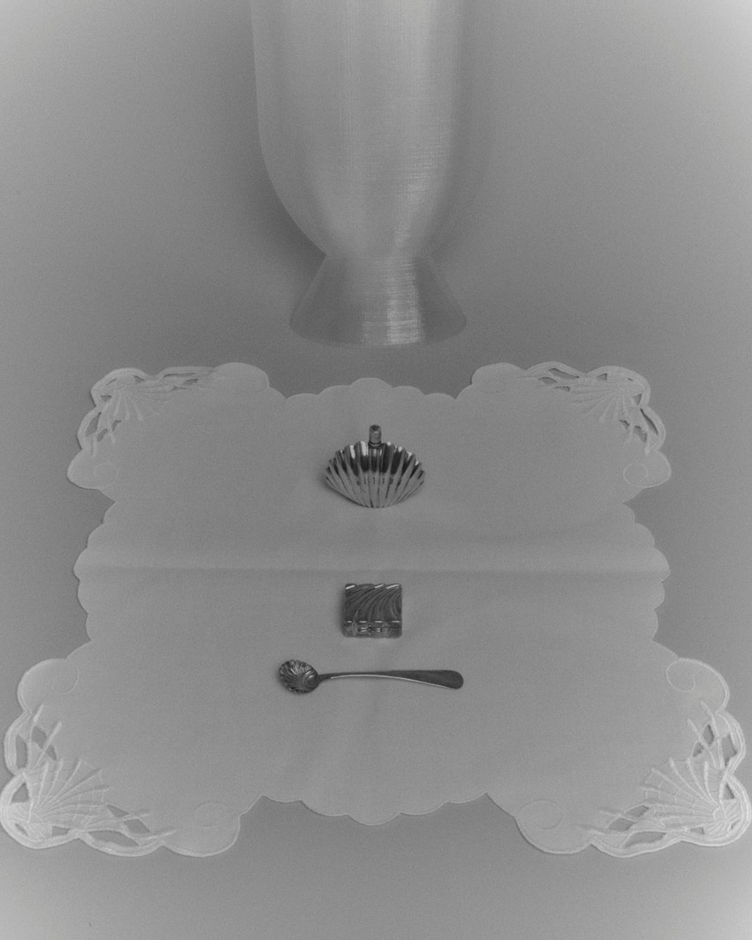 Still life shot of a silver scallop shell, embossed sugar cube, and ornate spoon on a white doily with an hourglass shape above.