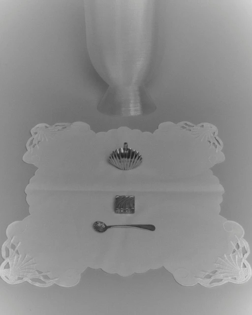 Still life shot of a silver scallop shell, embossed sugar cube, and ornate spoon on a white doily with an hourglass shape above.