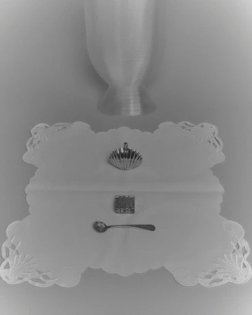 Still life shot of a silver scallop shell, embossed sugar cube, and ornate spoon on a white doily with an hourglass shape above.