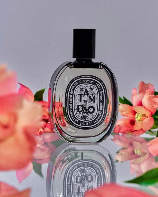 Elegant product shot of Diptyque Tam Dao perfume bottle with pink flowers and studio lighting on a reflective surface.