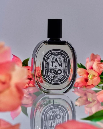 Elegant product shot of Diptyque Tam Dao perfume bottle with pink flowers and studio lighting on a reflective surface.