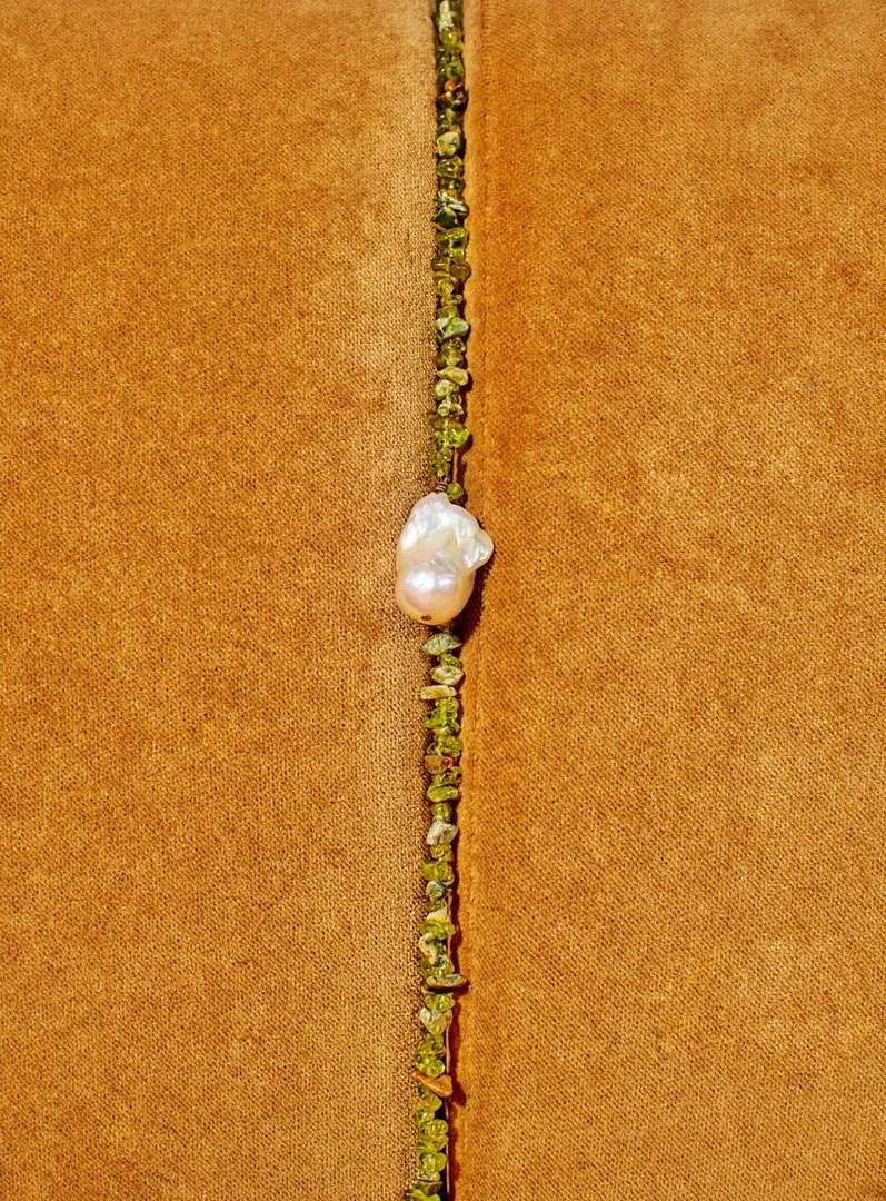 Macro photograph of a baroque pearl on a thin necklace with small green stones, set against a textured gold-toned textile background.