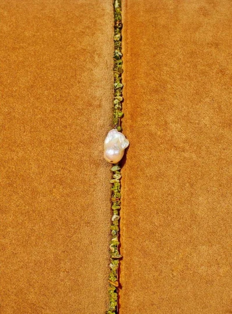 Macro photograph of a baroque pearl on a thin necklace with small green stones, set against a textured gold-toned textile background.