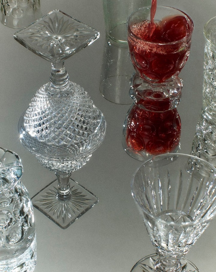 Elegant arrangement of stemmed crystal glasses and a tumbler with red liquid being poured, resting on a glossy, light-reflecting surface.