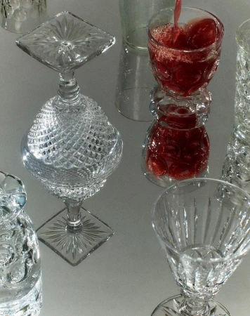 Elegant arrangement of stemmed crystal glasses and a tumbler with red liquid being poured, resting on a glossy, light-reflecting surface.