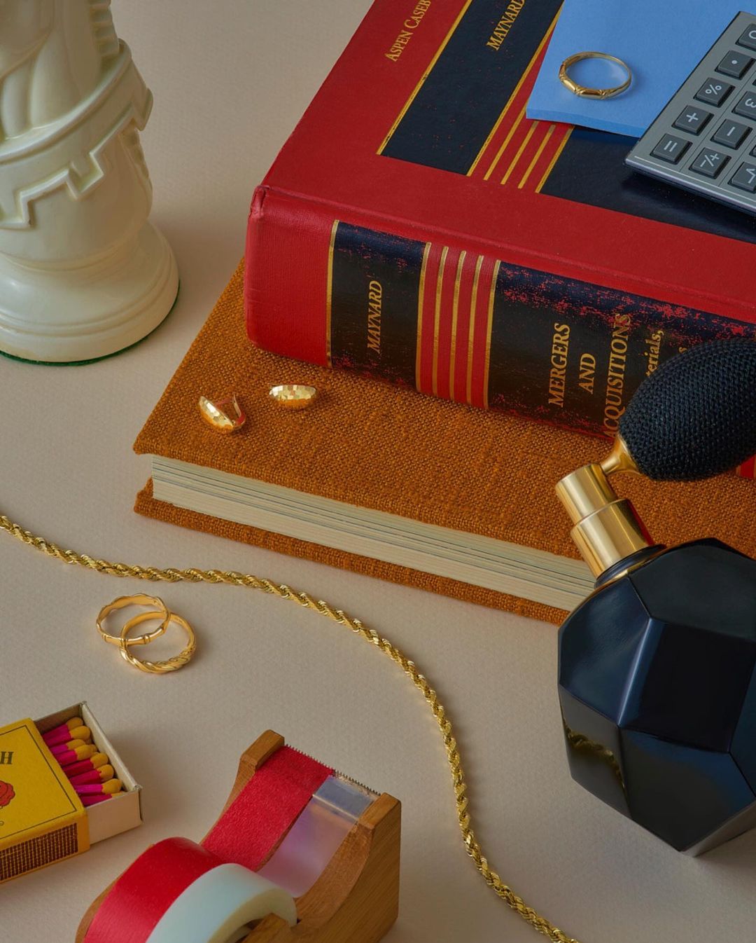 Still life of gold rings, books, matches, and perfume on a beige surface, creating a warm and luxurious visual composition.