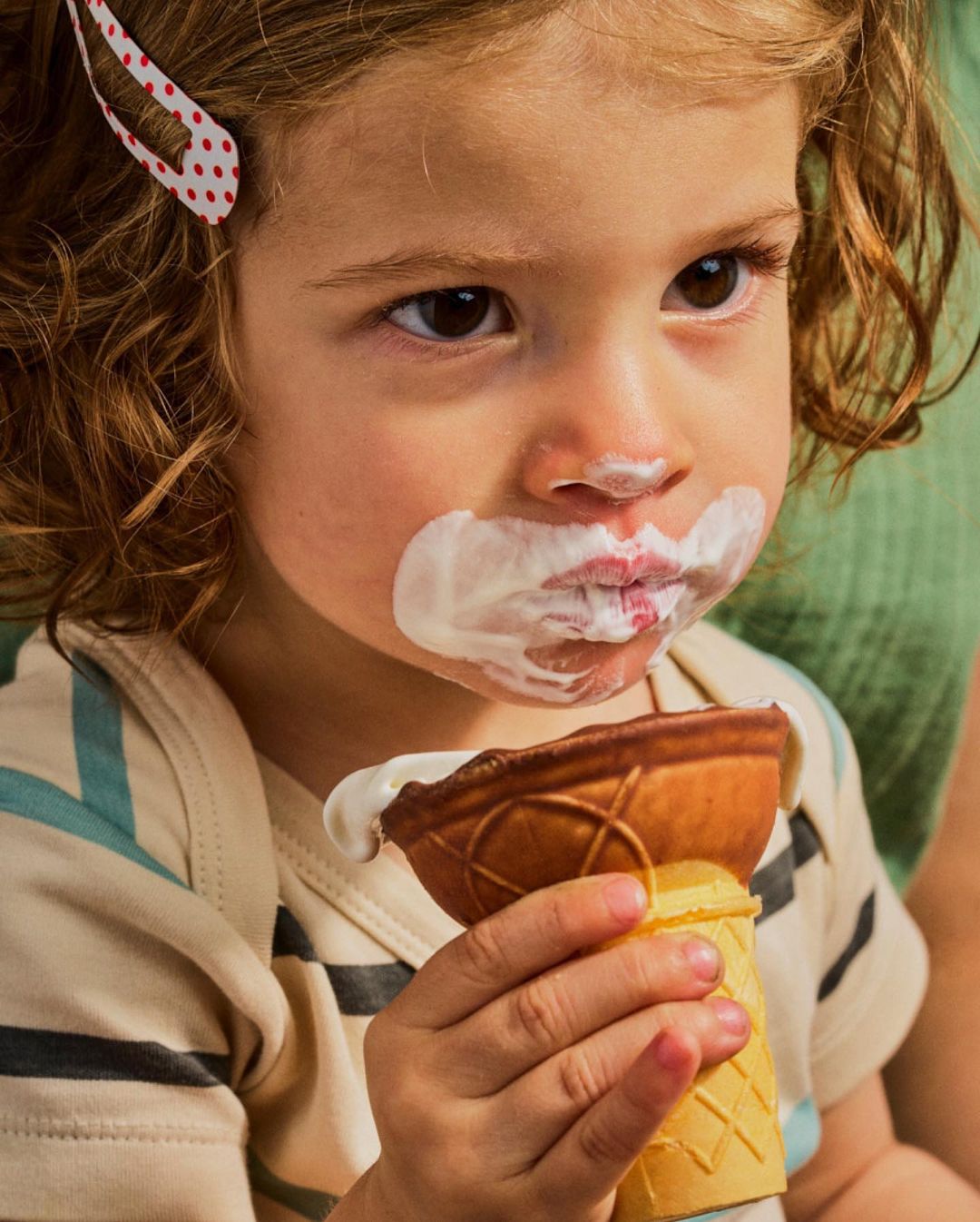 Toddler girl with an ice cream cone and white smeared on her face is enjoying a snack in an indoor setting.