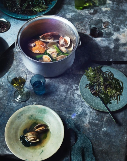Overhead shot of clam and fish stew in an aluminum pot surrounded by plates of seaweed, glasses, and dark cloth on a textured gray surface.