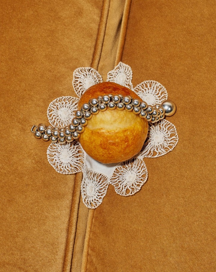 Close-up of bread roll on white lace doily, adorned with silver beaded necklace, set against brown suede-like material.