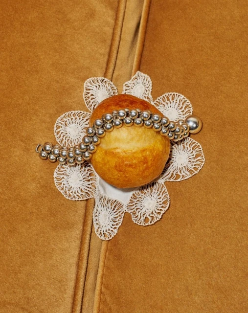 Close-up of bread roll on white lace doily, adorned with silver beaded necklace, set against brown suede-like material.