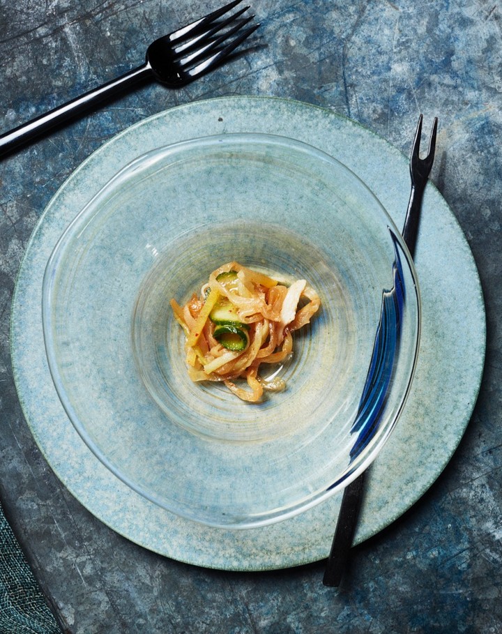 Overhead shot of a clear bowl containing food on a teal plate with black forks, set against a textured dark blue background.