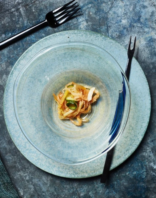 Overhead shot of a clear bowl containing food on a teal plate with black forks, set against a textured dark blue background.