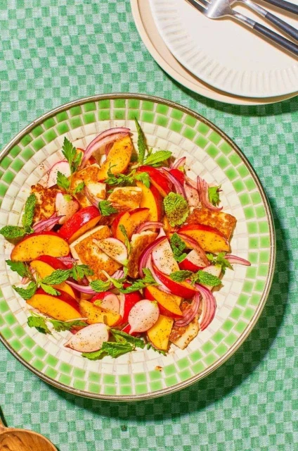 Overhead shot of peach and halloumi salad with mint and red onion on a decorative plate, atop a green checkered tablecloth.
