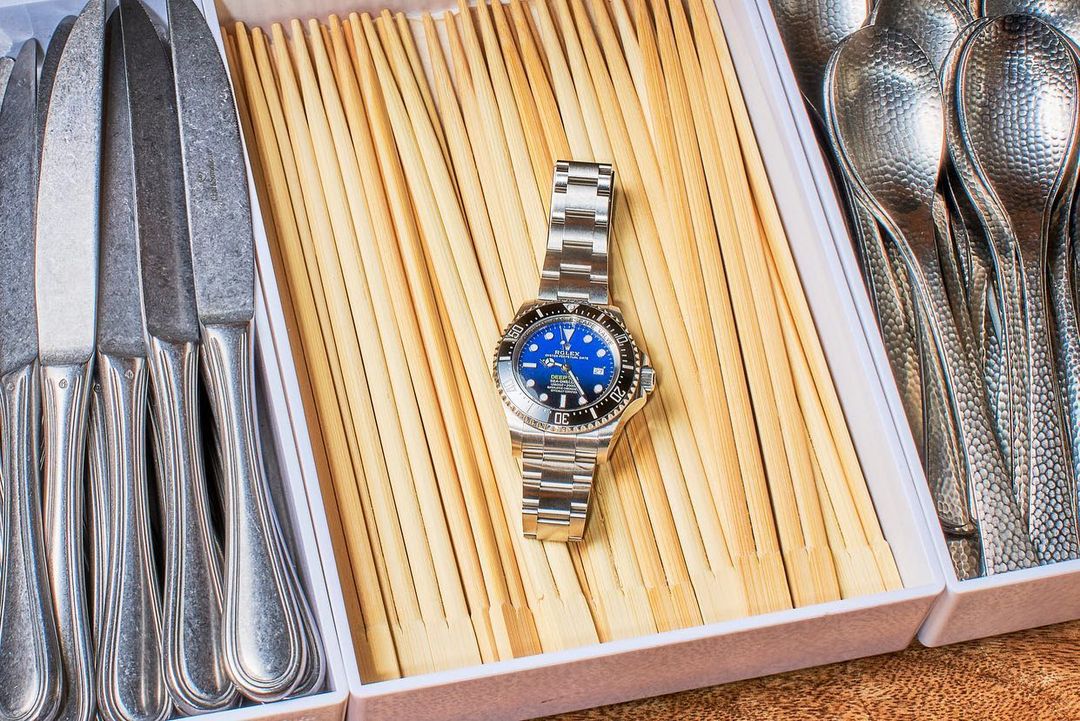 Close-up of a luxury wristwatch with a blue dial on a bed of wooden sticks, nestled among kitchen knives and spoons in a white tray.
