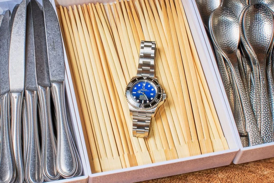 Close-up of a luxury wristwatch with a blue dial on a bed of wooden sticks, nestled among kitchen knives and spoons in a white tray.
