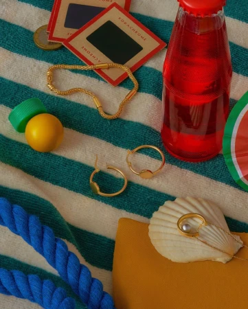 Still life of jewelry, a beverage bottle, and film slides on a striped beach towel with bright summer colors and a playful mood.