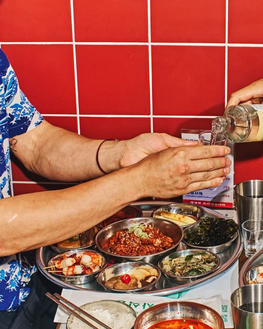 Korean meal featuring various side dishes served on a tray alongside a person pouring a drink into a glass in an indoor setting.
