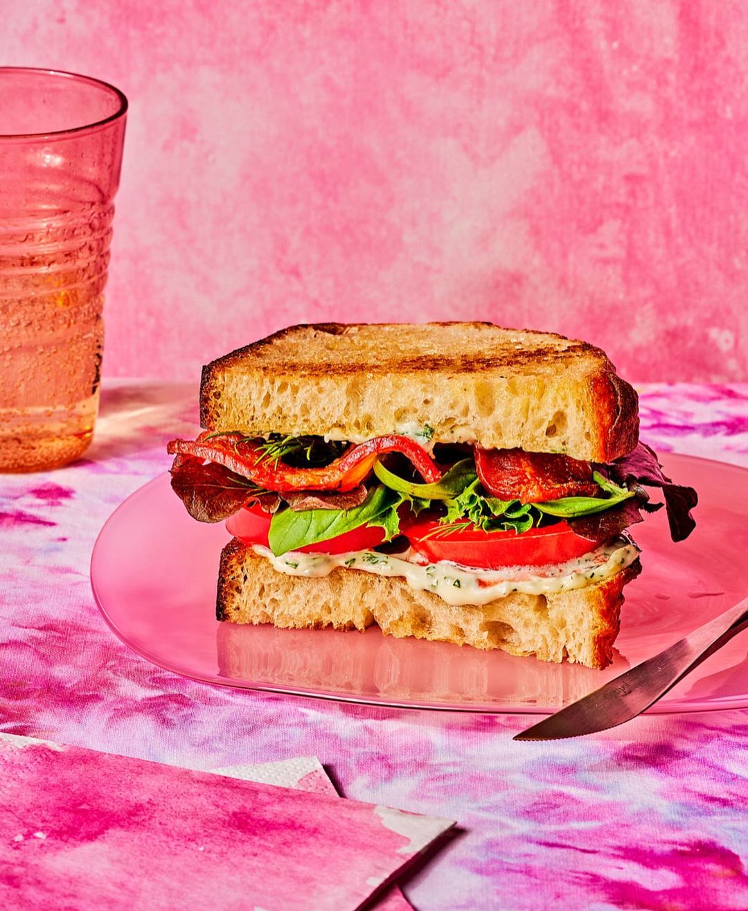 Grilled sandwich with sliced tomato, greens, and bacon on a pink plate next to a knife and glass, set against a pink background.