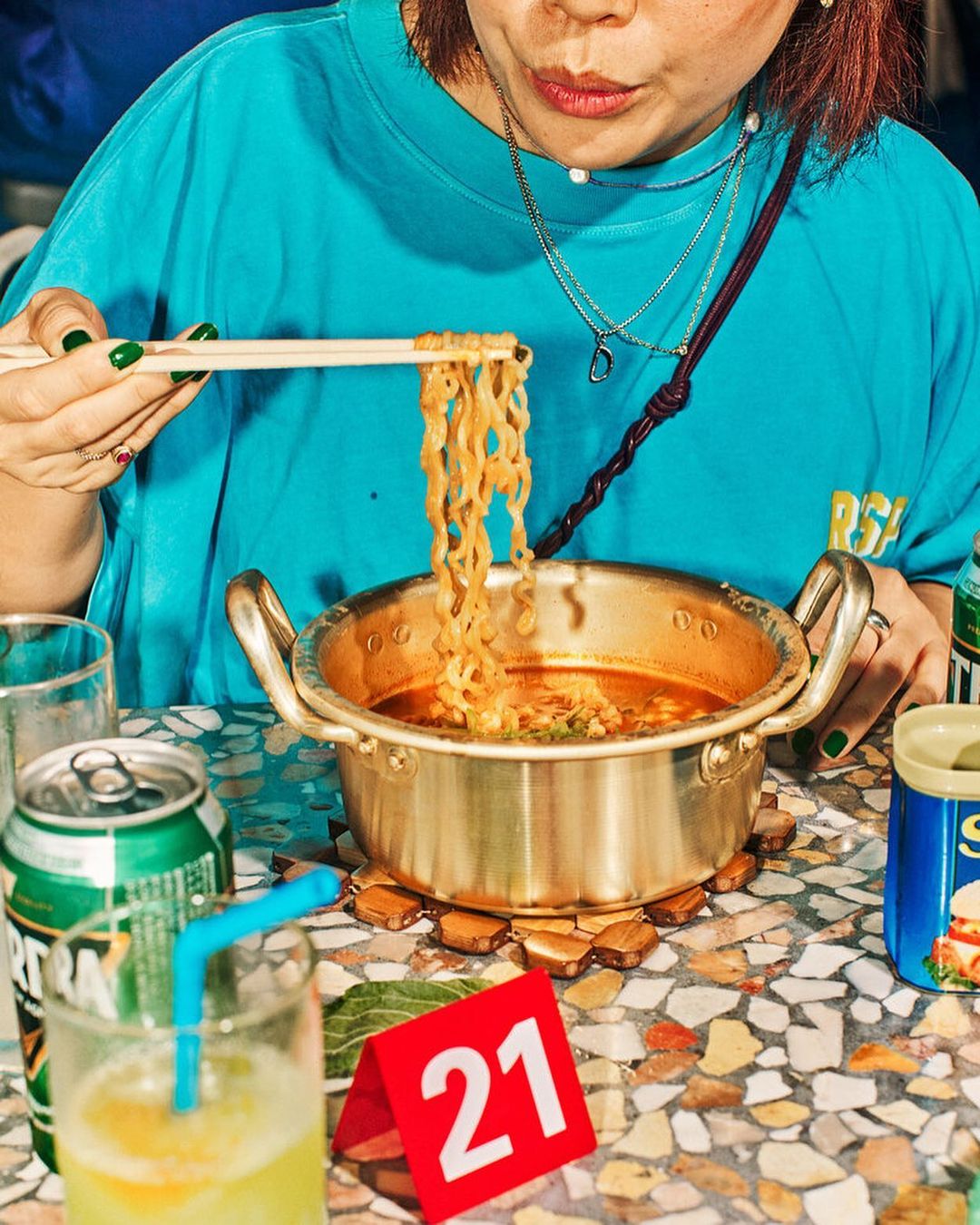 An individual is shown in a close-up shot while enjoying ramen with chopsticks, set against a backdrop of drinks and patterned table.