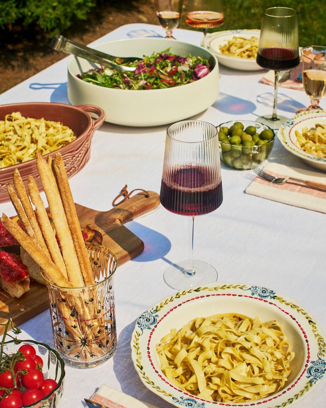 Outdoor table setting with pasta, salad, wine, breadsticks, and tomatoes, arranged on a white tablecloth with greenery visible.