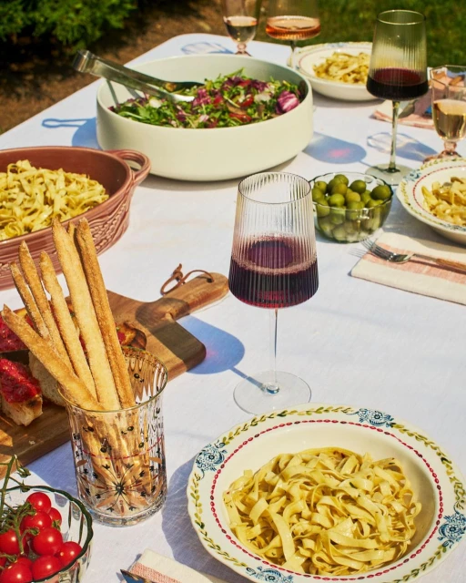 Outdoor table setting with pasta, salad, wine, breadsticks, and tomatoes, arranged on a white tablecloth with greenery visible.