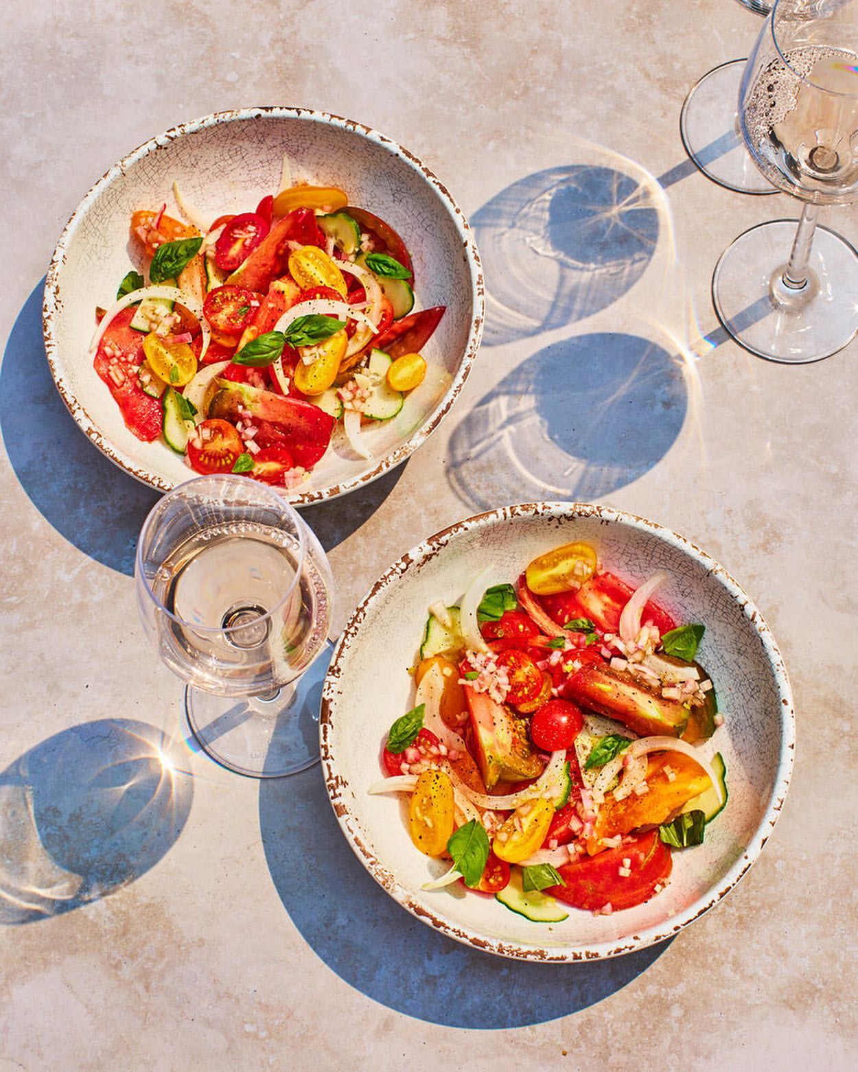 Two bowls of tomato salad with white wine served on a table in bright, natural light featuring tomatoes, cucumbers, and herbs.