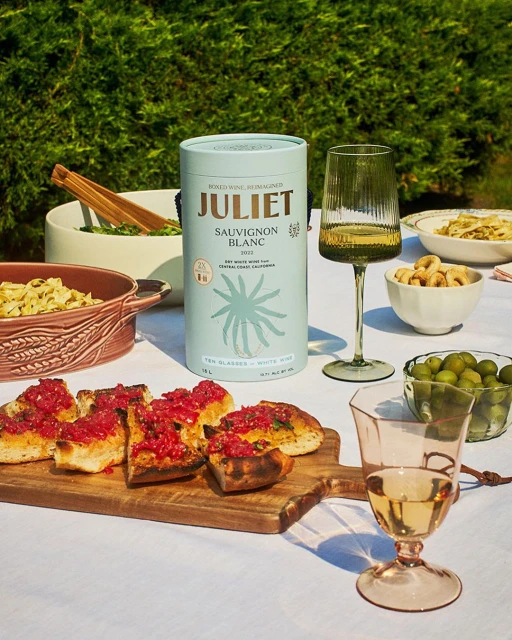 Still life with a light-blue can of Sauvignon Blanc, filled wine glasses, and Italian appetizers on a white tablecloth, with a green hedge background.
