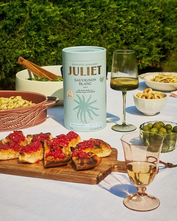 Still life with a light-blue can of Sauvignon Blanc, filled wine glasses, and Italian appetizers on a white tablecloth, with a green hedge background.