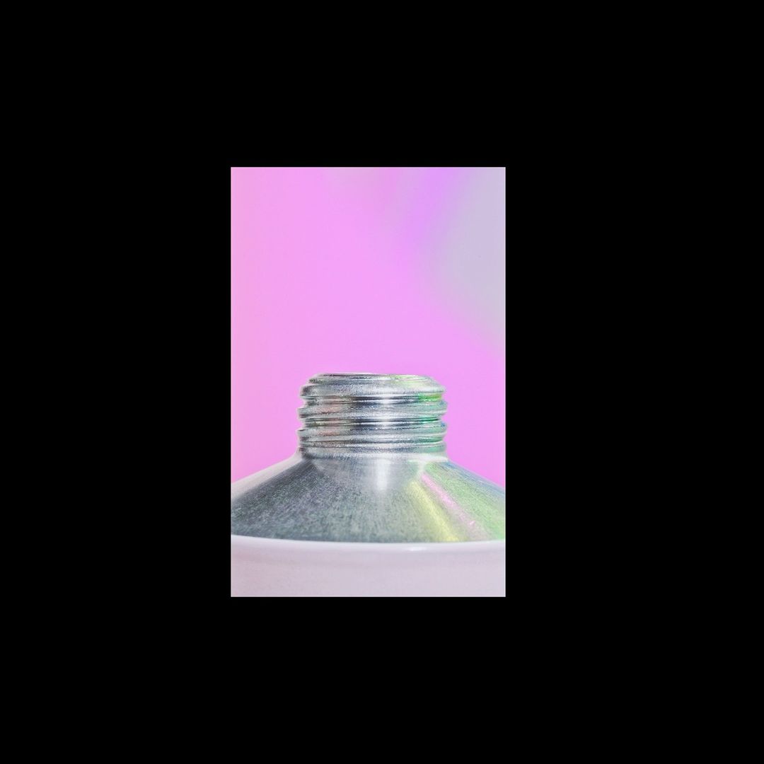 Close-up shot of a silver metal tube against a soft pink background, showcasing its ridged neck and white base, isolated against a black backdrop.