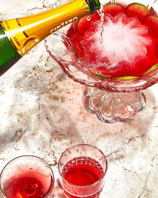 Champagne being poured into a punch bowl filled with a red drink and lime slices, with two glasses filled with the same drink.