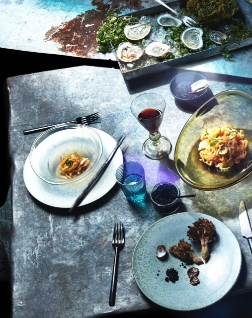 Overhead shot of a table featuring oysters, ceviche, mushrooms, and caviar, alongside wine, water, forks, and knives, set against a textured gray surface.