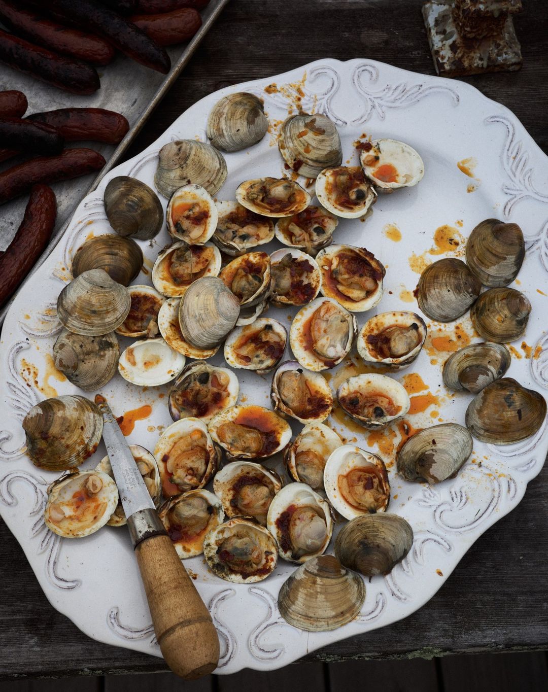 Overhead shot on a white decorative platter of cooked clams with dark brown edges and red sauce, alongside grilled hotdogs and a knife.