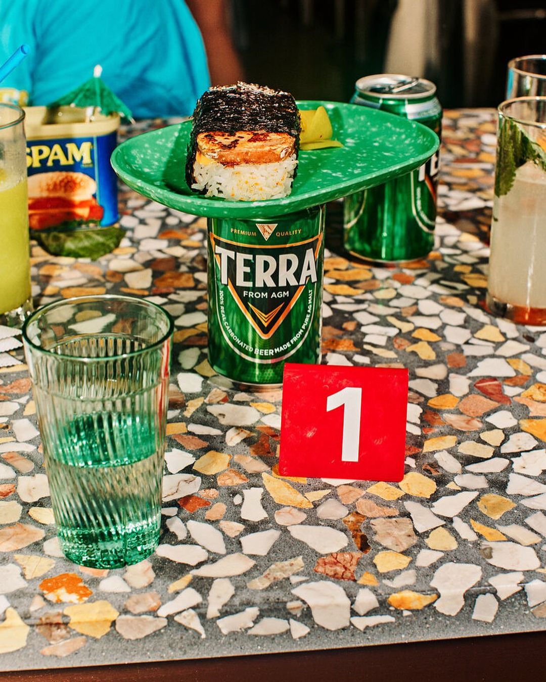 Food on a green plate elevated by a beer can on a speckled table, featuring drinks, Spam, and table number '1' in an indoor setting.