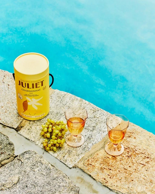 Still life near pool: yellow wine cylinder with wine glasses, green grapes on stone surface, with vibrant turquoise water backdrop.