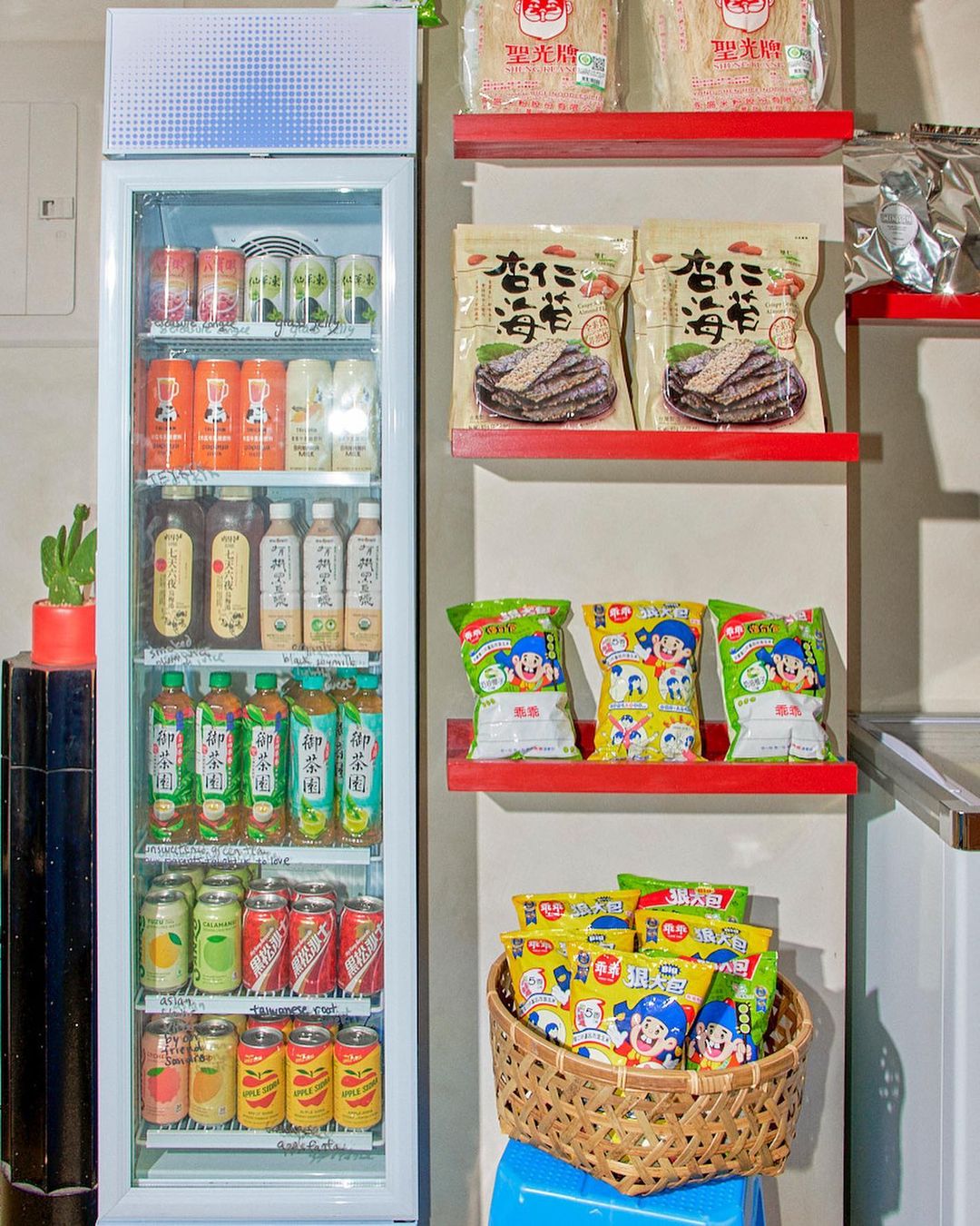 Well-stocked beverage refrigerator with soft drinks, tea, and snacks; chips are displayed on red shelves and in a woven basket.