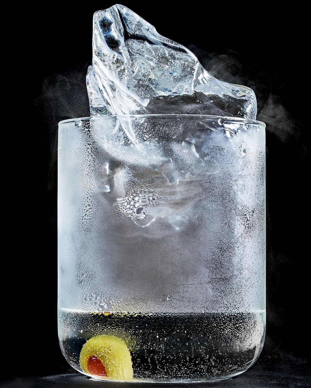 Close-up of clear glass filled with clear liquid, a large ice cube, and a green olive with pimento, set against a solid black background.