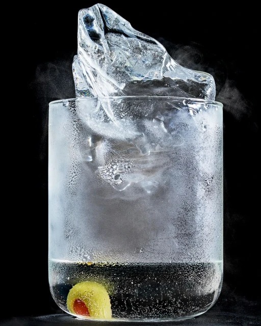 Close-up of clear glass filled with clear liquid, a large ice cube, and a green olive with pimento, set against a solid black background.