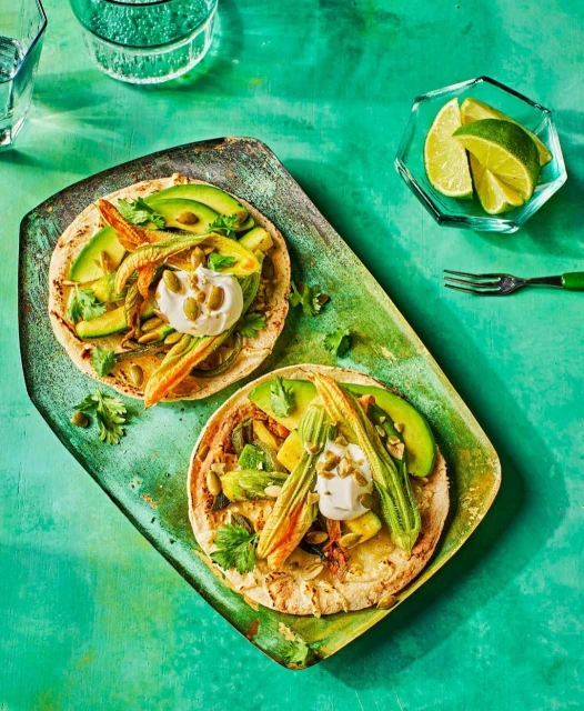 Overhead shot of two tostadas topped with avocado slices, squash blossoms, and crema on a rustic green serving board with limes.