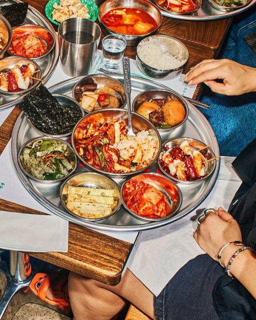 Overhead shot of a silver tray filled with small bowls of colorful Korean side dishes (banchan) served with eating utensils.