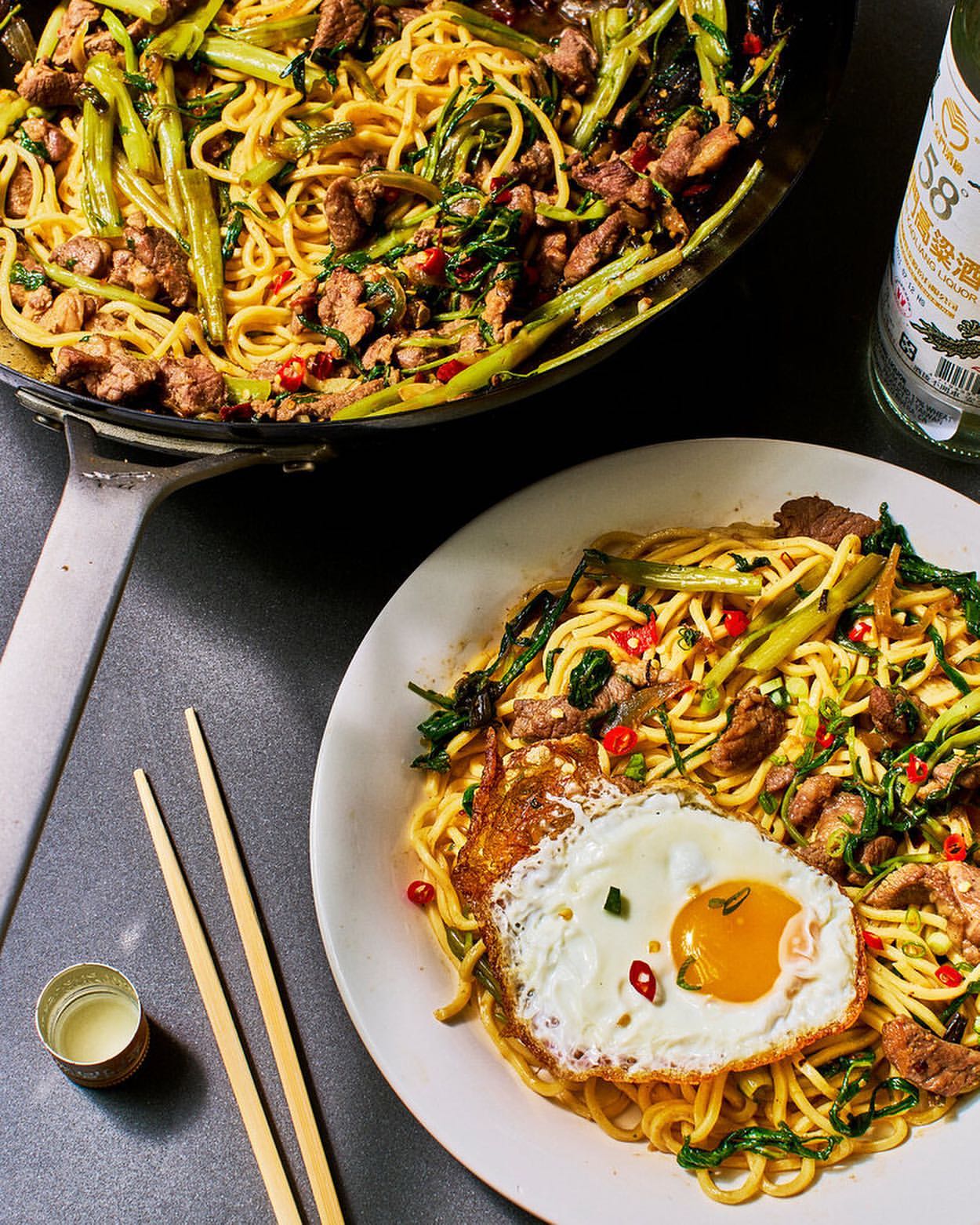 Overhead shot of a plate with noodles, a fried egg, and a wok filled with more noodles and ingredients next to bottle and chopsticks.