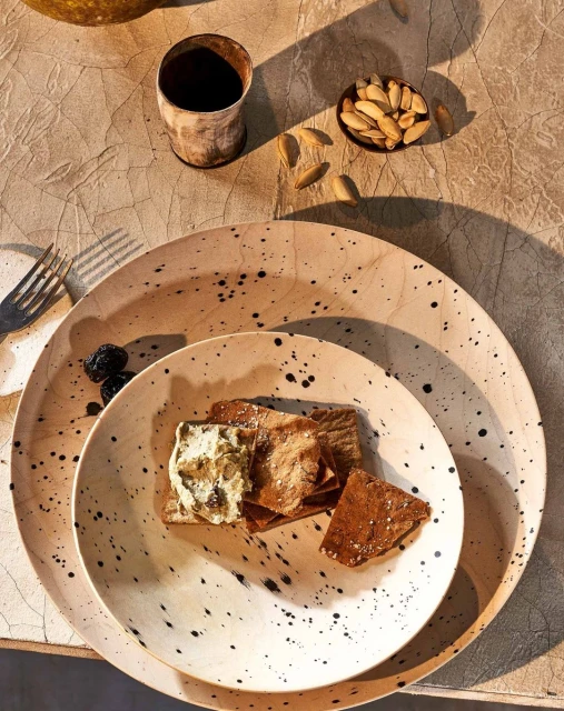 Still life of food on plates, featuring crackers topped with a spread, nuts in a bowl, and a drink in a rustic setting with neutral tones.