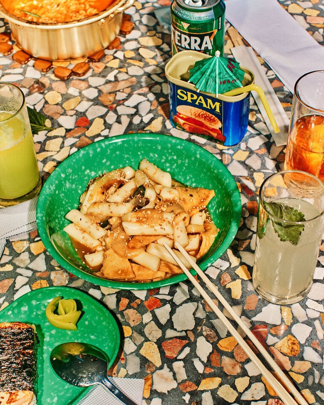 Overhead shot of a tabletop with bowls of rice cakes, drinks, and canned meat with a terrazzo surface and bright, appetizing presentation.