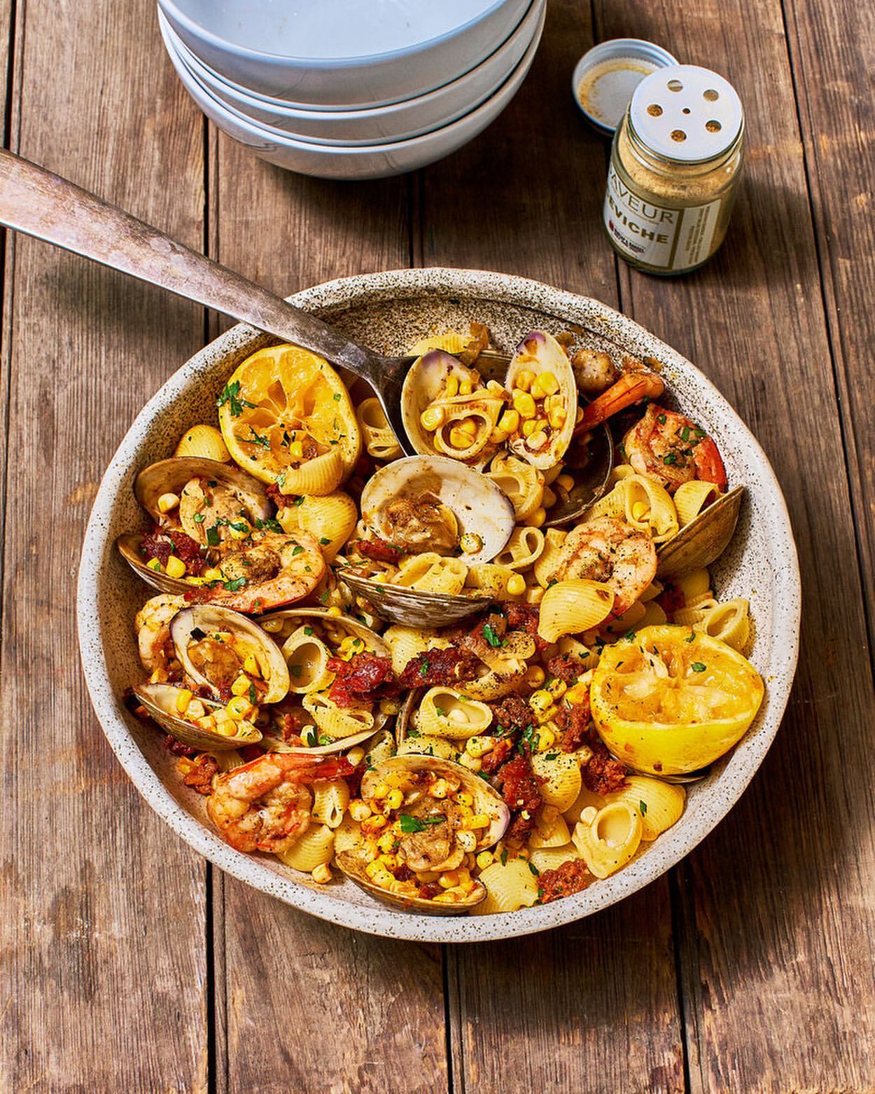 Overhead shot of pasta with clams, shrimp, corn, and lemon in a ceramic bowl with a spoon on a wooden table with plates.