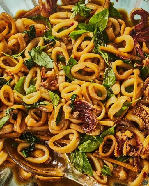 Overhead close-up of stir-fried squid rings with basil leaves and a light sauce in a bowl, showcasing textures and colors.