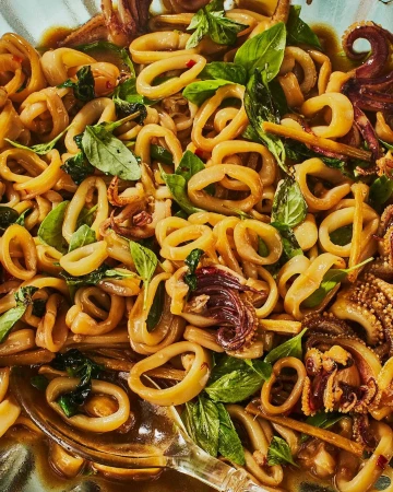Overhead close-up of stir-fried squid rings with basil leaves and a light sauce in a bowl, showcasing textures and colors.