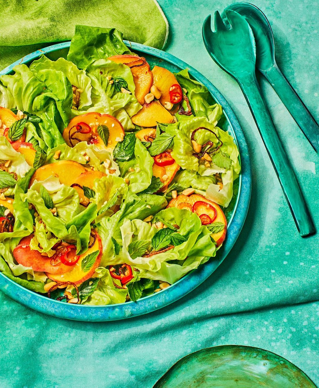Overhead shot of a vibrant salad featuring lettuce, sliced peaches, mint, and other toppings in a blue bowl, next to green serving utensils.