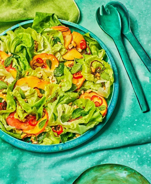 Overhead shot of a vibrant salad featuring lettuce, sliced peaches, mint, and other toppings in a blue bowl, next to green serving utensils.