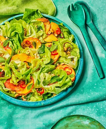 Overhead shot of a vibrant salad featuring lettuce, sliced peaches, mint, and other toppings in a blue bowl, next to green serving utensils.